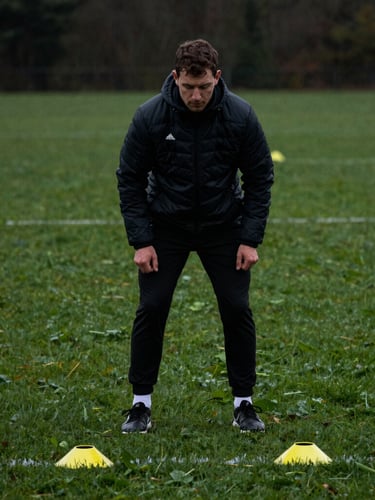 Male football coach setting up agility cones on dew-covered grass under overcast daylight, deep charcoal and grass-green tones, shallow depth of field, moody athletic photography.