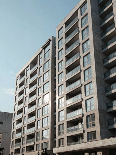 Wide architectural exterior of a mid-rise urban residential building, shot from street level in sharp morning daylight, concrete and glass facade filling the right two-thirds of frame, clean sky on the left, no people, no staging