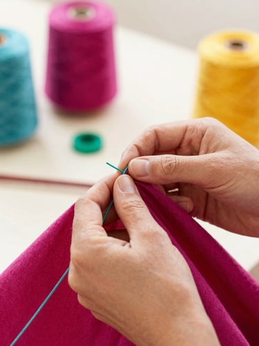 Hands of an artisan carefully stitching a colorful magenta fabric with teal threads in a bright, warm workshop in Celaya, soft focus on the needlework, warm daylight