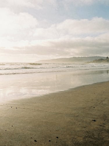 A quiet, overcast morning on a Southern California coast, soft light reflecting on wet sand at low tide, environmental, 35mm film grain.