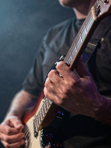 Tight close-up of a guitarist's fretting hand on the neck of an electric guitar mid-performance, fingers pressing strings, stage light from the right casting hard shadows across knuckles, background a dark blur of stage haze and crowd