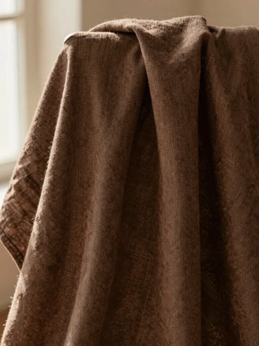 Close-up of high-quality brown linen fabric texture draped softly over a wooden chair, warm natural window light, editorial fashion setting