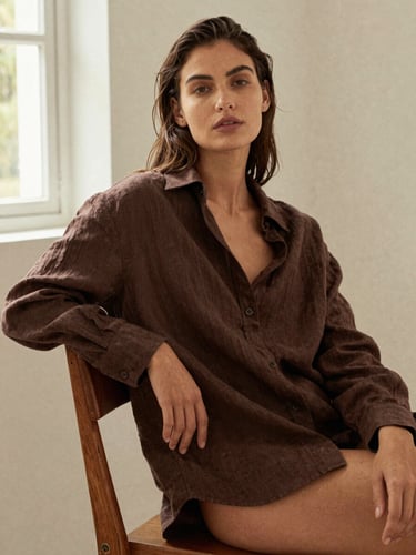 Warm editorial fashion photography, a woman in a relaxed pose wearing a drape-heavy brown linen shirt, sitting on a wooden chair against a soft off-white wall, natural window light, 35mm film.