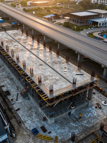 Wide aerial drone shot of a massive highway concrete foundation under construction at golden hour, showing scale and steel reinforcements.