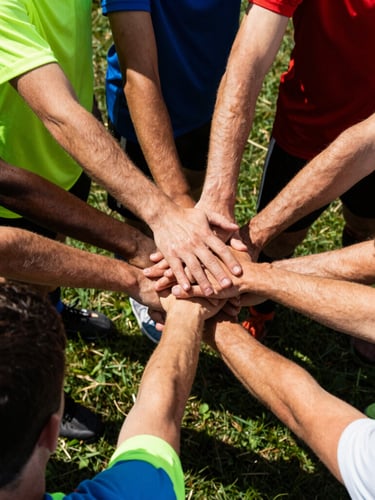 Close-up of a diverse group of athletes in high-contrast late-afternoon sun, hands stacked together in a team huddle on a grass field, vibrant athletic jerseys.