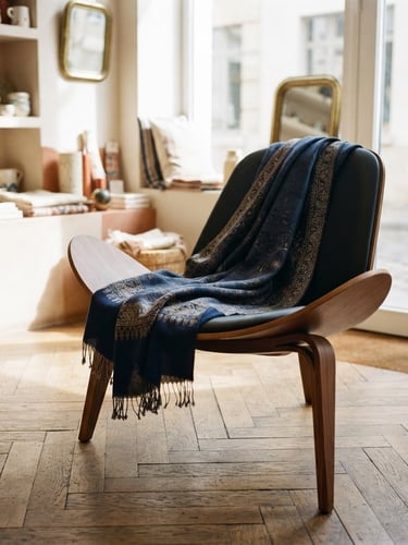 A high-end editorial shot of a luxury Kashmir Pashmina shawl elegantly draped over a minimalist modern chair in a sunlit European boutique, natural directional light, rich indigo accents, sharp focus.