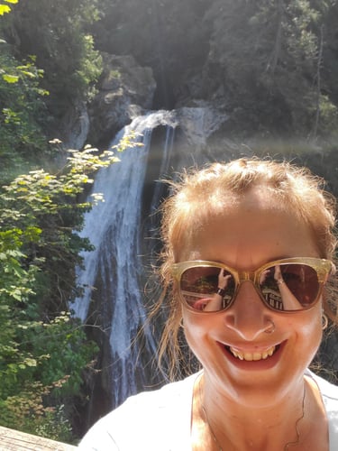Smiling woman with sunglasses taking a selfie in front of a scenic mountain waterfall.