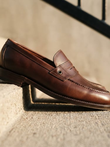 Editorial fashion photography, low-angle shot of a luxurious leather loafer on a minimalist concrete step, warm natural sunlight, sharp focus on hand-stitched details