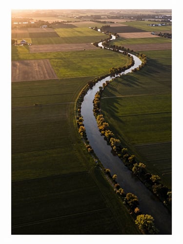 Aerial still photograph from drone altitude looking down over a patchwork of agricultural fields and a winding river at golden hour, precise tonal range, natural light, no people, landscape geometry clearly defined