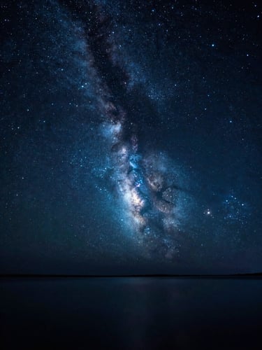 Wide-angle starry night sky reflecting on calm dark water, glowing nebulae in deep indigo and teal, soft luminous starlight, cinematic photography