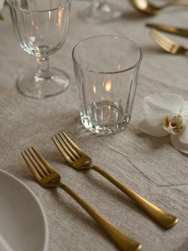 Close-up of a bespoke table setting, textured neutral linen, fluted glassware reflecting low-contrast warm light, brushed brass cutlery, single white orchid.