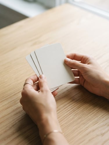 Close-up of hands holding clean therapy cards on a light wooden table, soft natural daylight, muted sophisticated tones