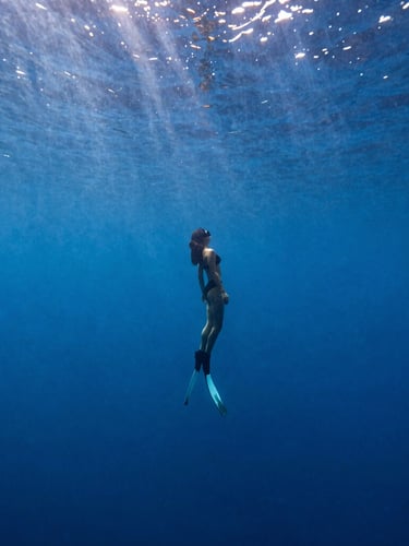 A lone freediver descending vertically into deep blue ocean water, natural sun rays filtering from above, serene and minimalist underwater film photography style.