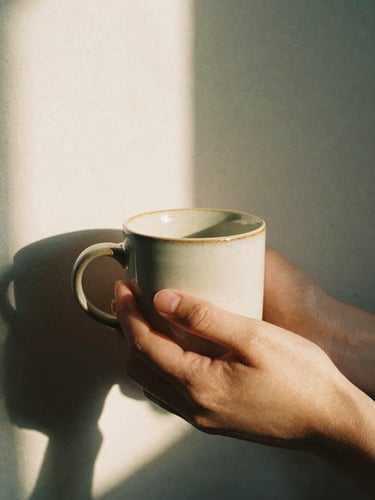 Close-up of hands holding a warm ceramic mug, soft late afternoon sunlight casting long shadows on a textured neutral wall, cozy and reflective atmosphere, 35mm film
