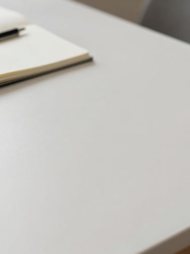 A clean, minimalist desk surface with soft natural daylight, showing a closed notebook and a pen resting on top, generous negative space, calm and organized composition.