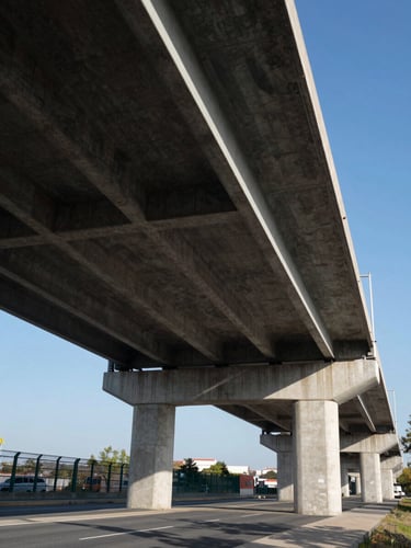 Wide-angle ground-level view of a completed steel-and-concrete highway overpass under sharp midday sun, geometric shadow patterns cast across the underside of the deck, clear blue sky, no people