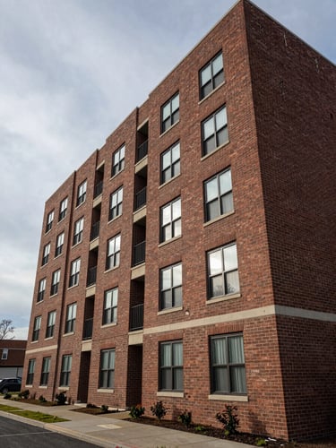 Wide-angle exterior of a completed mid-rise apartment building, six to eight stories, photographed from street level on an overcast morning—natural diffused light reveals brick facade texture and window-frame joins cleanly; building occupies the right two-thirds of the frame, generous sky and sidewalk context on the left
