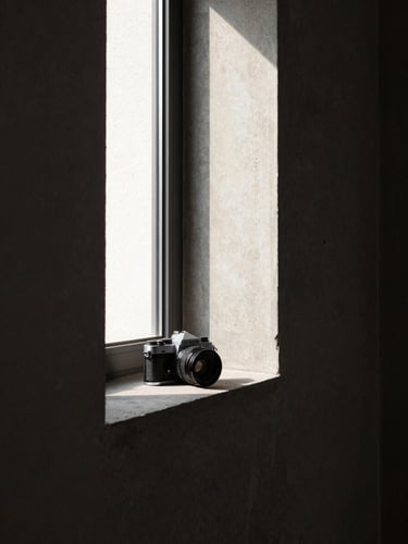 Narrow window frame casting a sharp rectangle of afternoon light across a concrete wall, a 35mm film camera placed at the edge of the light band, architectural precision, high contrast, no people