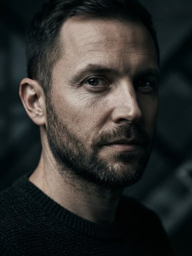 Close-up macro of a minimalist author portrait in low-key studio lighting, deep charcoal shadows, sharp contrast, professional editorial look, looking slightly off-camera.