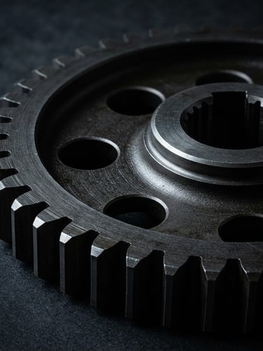 Dramatic studio product photography, extreme close-up of a billet-machined steel gear with sharp metallic rim-lights against a dark charcoal background, 85mm lens