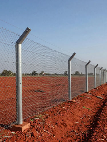 Wide-angle shot of a long, straight galvanized chain-link fence stretching across a red soil agricultural field in Gujarat, bright midday sun reflecting off the metallic wire, concrete RCC poles spaced evenly, clear blue sky.