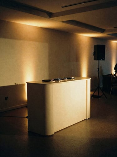 Sleek, wire-free DJ booth in a modern minimalist banquet hall, warm golden uplighting accenting the clean lines, moody shadows, 35mm film grain