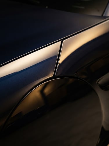 Extreme close-up of a flawless dark car fender, showing deep, liquid-like reflections under high-contrast studio lighting, sharp body lines, warm gold and dark navy tones, 85mm.