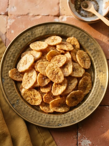 Overhead shot of golden-brown banana chips resting on a traditional brass plate, warm golden-hour sunlight casting soft shadows, 35mm editorial food photography