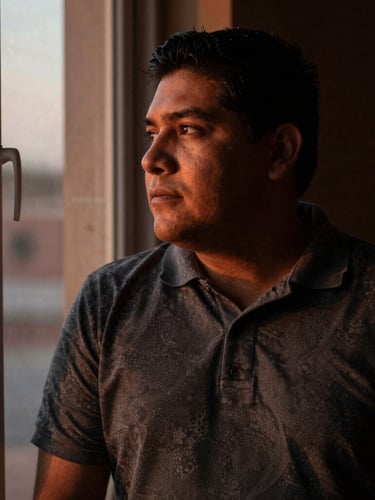 Environmental portrait of a Latino man in quiet reflection near a window, soft twilight shadows, warm bronze and charcoal tones, intimate depth of field