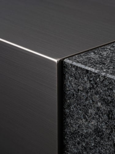 Macro photograph of a dark brushed steel surface meeting a textured charcoal marble slab at a precise, razor-sharp geometric angle, soft museum-grade side lighting, high-contrast shadows.