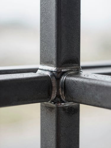 Extreme close-up of a custom steel window grill, tight frame on the welded intersection joints, bright overcast light revealing the surface texture and weld bead quality, sharp depth of field on the connection points, charcoal-gray steel finish