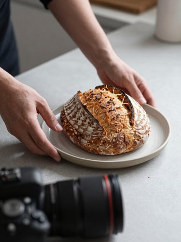 Close-up of hands adjusting a ceramic plate with artisanal sourdough bread under soft natural light, a professional camera lens visible in the foreground, Scandinavian minimalist kitchen setting.
