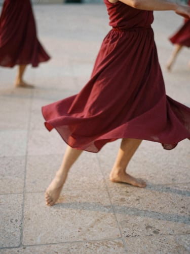 Moody 35mm film photograph of dancers in motion blur on a textured stone plaza, warm natural sunset light, deep cherry red fabrics flowing, organic and raw atmosphere