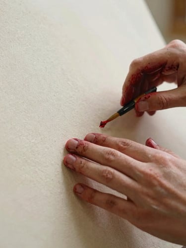 Close-up of hands covered in charcoal and red paint on a raw textured canvas, soft natural indoor lighting, warm cream tones, shallow depth of field