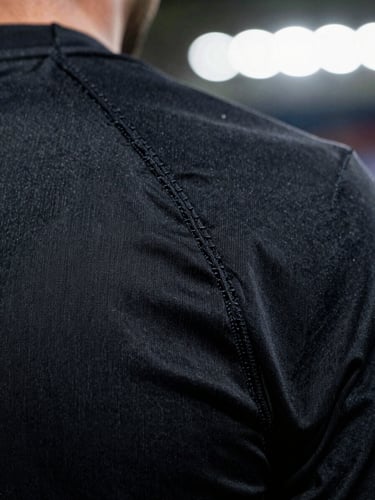 Extreme macro close-up of high-tension flatlock stitching on a matte black athletic jersey, dramatic stadium rim lighting, sharp focus on technical fabric fibers.