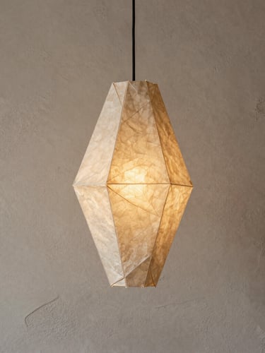 A tall portrait shot of a single handcrafted origami pendant lamp made of textured beige paper, unlit, hanging against a raw plaster wall in soft morning side light.