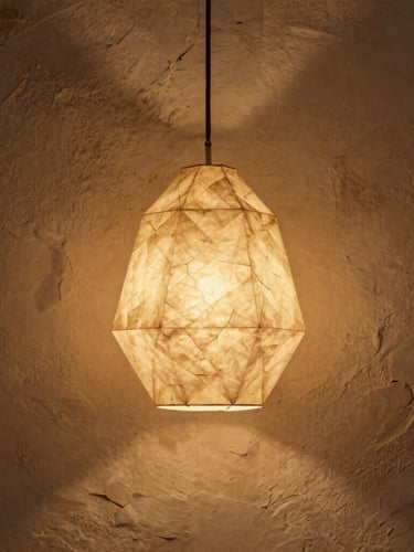 Portrait-oriented shot of a geometric origami pendant lamp made of textured beige paper, glowing with warm internal light, casting soft shadows on a rustic plaster wall, Saint-Paul-de-Vence atelier style, golden hour.