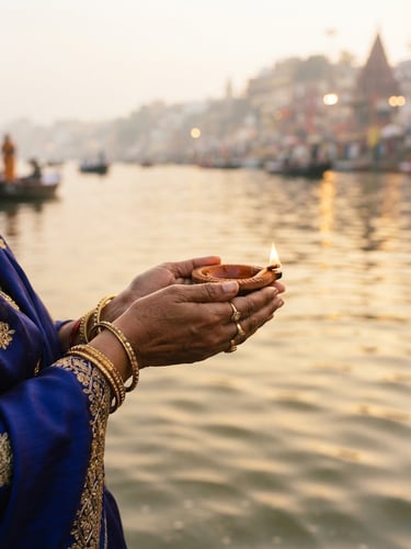 A close-up photograph of hands holding a small, traditional clay diya with a warm glowing flame, soft morning light reflecting off calm river water, shallow depth of field.