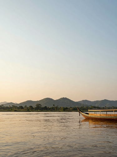 Wide landscape framing of the Mekong River at golden hour, a traditional wooden longtail boat moored at the right edge of frame, warm amber light raking across still water, lush green jungle hills in the far background, generous open sky occupying upper two-thirds