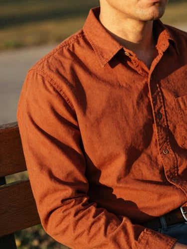 A close-up of a man in a warm terracotta heavyweight cotton overshirt, sitting on a wooden bench in golden-hour sunlight, looking relaxed, shallow depth of field, 35mm film.
