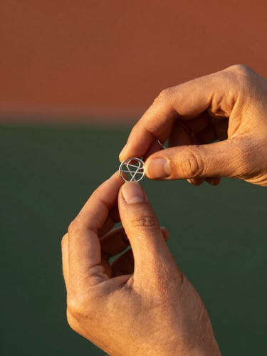 Close-up of hands measuring a delicate silver sacred geometry ring with a thread, warm golden-hour light, terracotta background, shallow depth of field