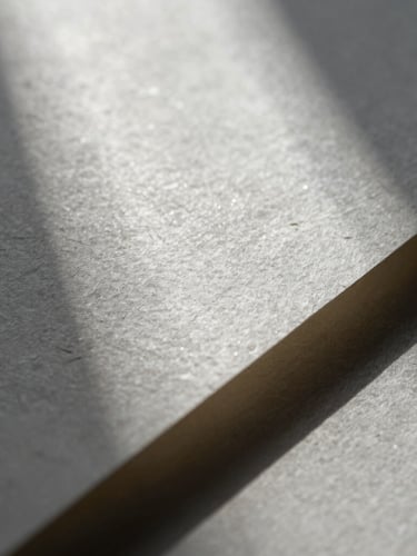 Macro close-up of textured handmade paper catching soft silver raking light, deep shadows, elegant and highly tactile.