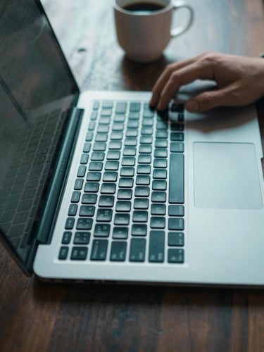 Close-up overhead of a laptop keyboard on a dark wood desk, screen casting a cool teal glow across the keys, a single hand resting mid-type, coffee mug blurred in the background — daylight from a side window crossing the left edge of the frame