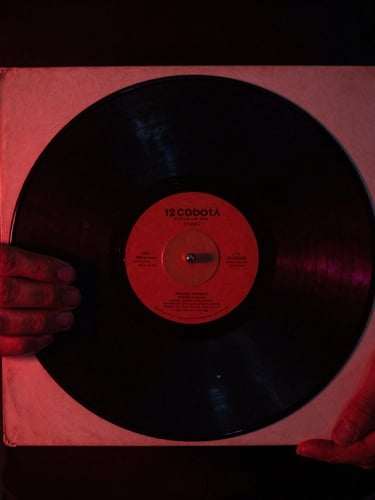 Moody close-up of hands holding a vintage 12-inch vinyl sleeve, dramatic low-key lighting, deep red undertones, 35mm film grain