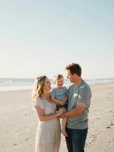 A candid lifestyle portrait of Hallie, her fiancé, and their young son laughing together on a sun-drenched Maine beach, soft pastel tones, shallow depth of field, 35mm lens, low horizon, expansive sky.