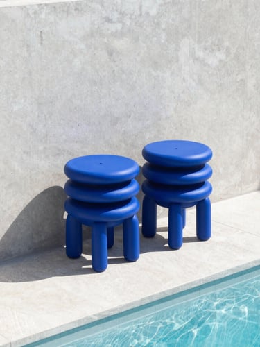 Cobalt blue monolithic resin stools stacked neatly against a sun-drenched concrete wall beside a sparkling pool, hard direct sunlight, sharp graphic shadows, architectural minimalism.