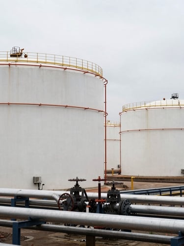 Wide environmental shot of massive industrial crude oil storage tanks in a terminal, overcast sky, heavy steel piping manifolds in the foreground, gritty industrial photography