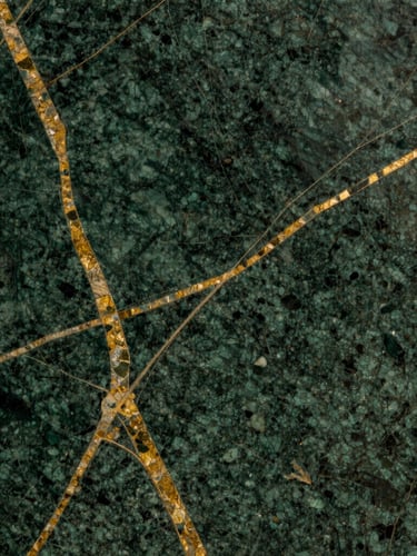 Macro close-up of premium dark green marble slab with natural gold veins, dramatic side lighting highlighting the polished texture, rich warm tones, editorial photography