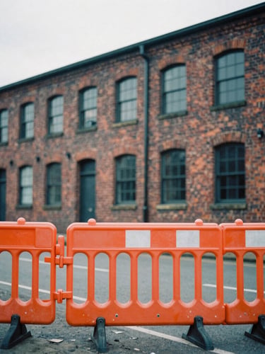 Gritty editorial photo of Salford brick industrial architecture, natural overcast daylight, desaturated cool tones with a sharp orange safety barrier in the foreground, 35mm film grain.