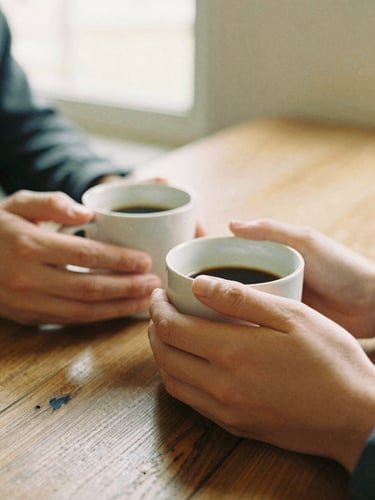 Close-up shot of two people holding warm mugs of coffee, hands resting on a rustic wooden table, soft focus background, warm golden hour light, 35mm film style.